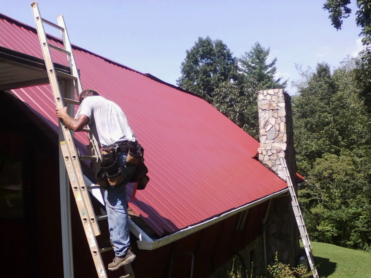 Roofer on a ladder beside a red metal roof and stone chimney — Soffit & Fascia Repair by Roofing Brothers in Dufur